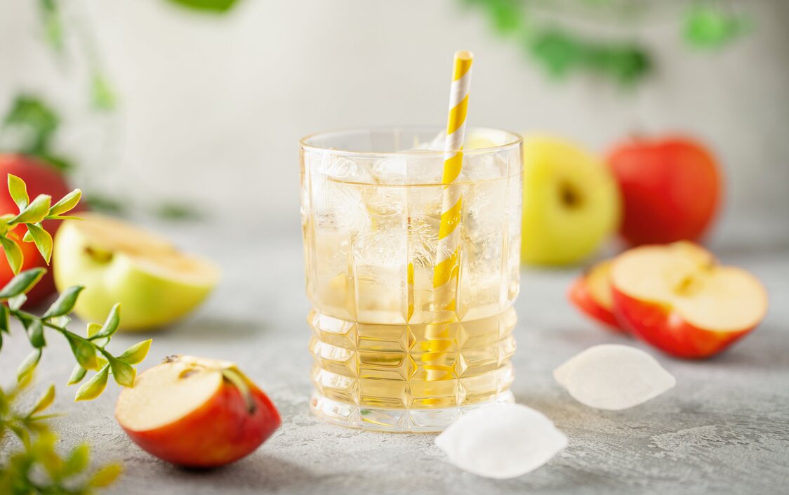 Ein Glas mit Apfelsaftschorle, Eiswürfel und ein Strohhalm und drum herum liegen Äpfel.