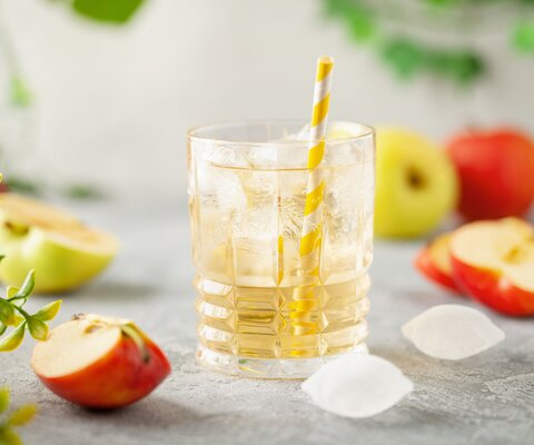 Ein Glas mit Apfelsaftschorle, Eiswürfel und ein Strohhalm und drum herum liegen Äpfel.