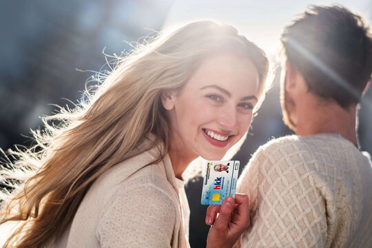 Ein junges Paar steht in der Stadt. Die Frau dreht sich zur Kamera und hält ihre Gesundheitskarte hin.