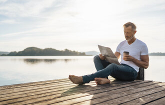 Ein Mann sitzt auf einem Steg am Wasser und liest Zeitung. Er hat einen Kaffee To-Go dabei.