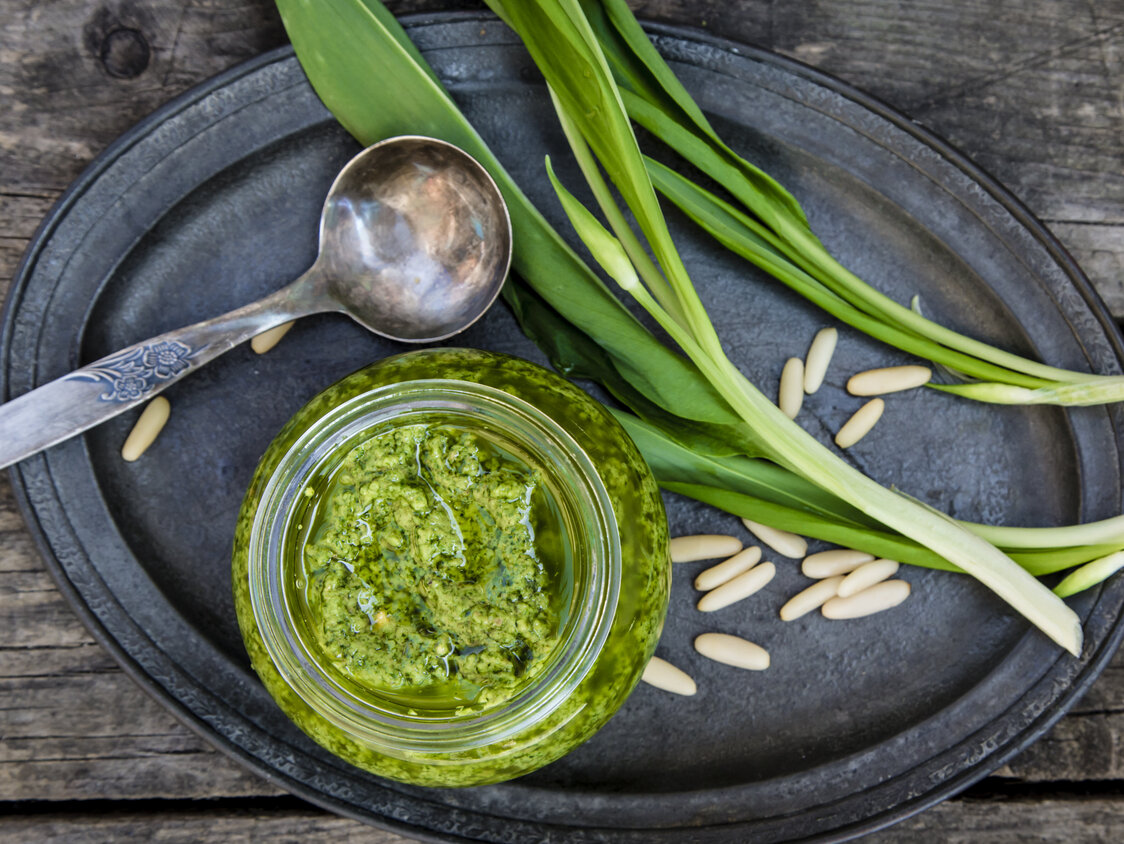 Frisches Bärlauch liegt neben einem Glas Pesto und einem Löffel auf einem ovalen Teller.