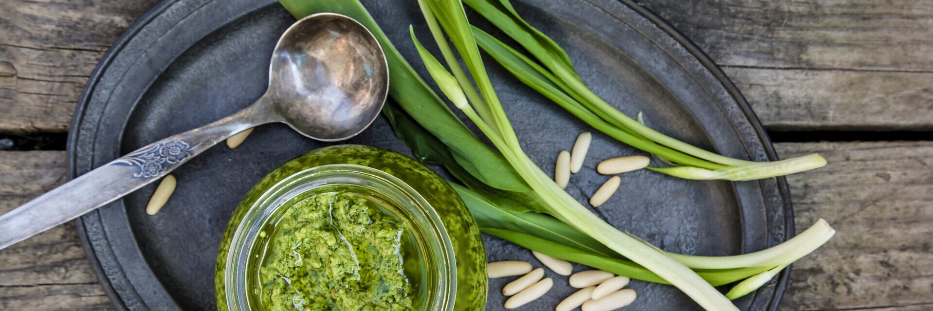 Frisches Bärlauch liegt neben einem Glas Pesto und einem Löffel auf einem ovalen Teller.
