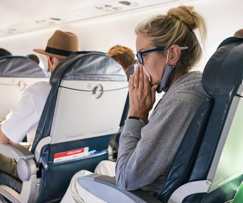 Frau mit Mundschutz sitzt in einem Flugzeug auf einem Platz und schnaubt sich Ihre Nase.