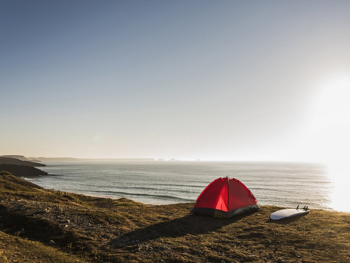 Ein rotes Zelt steht am Strand und ein Surfbrett liegt daneben während die Sonne untergeht.