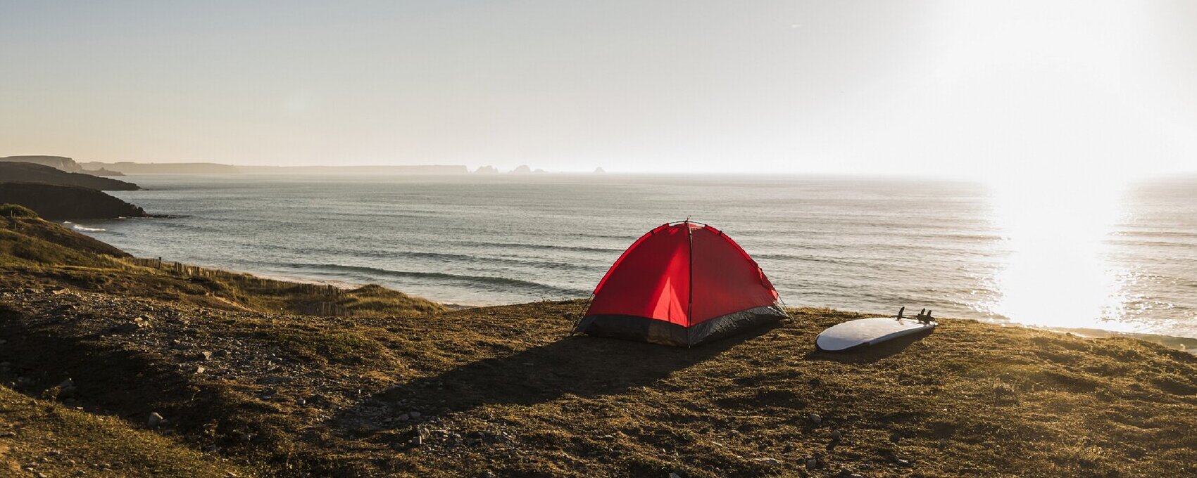 Ein rotes Zelt steht am Strand und ein Surfbrett liegt daneben während die Sonne untergeht.