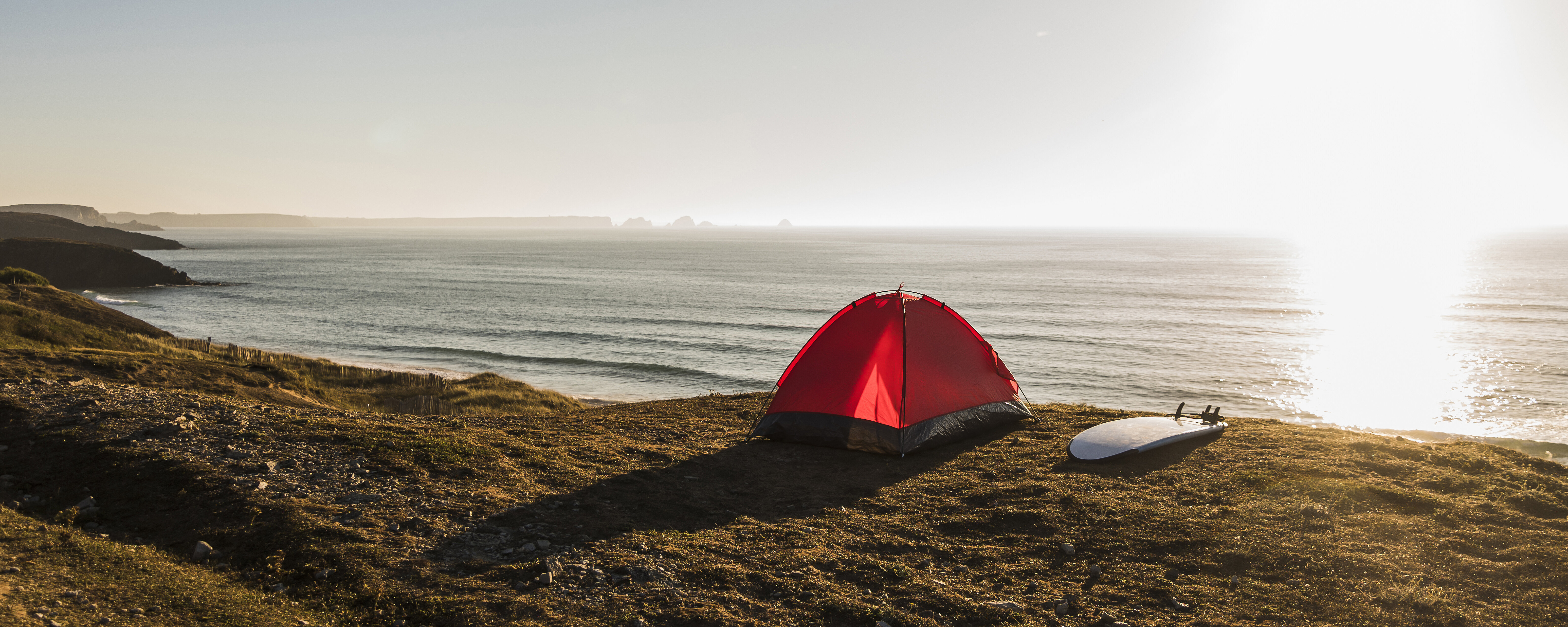 Ein rotes Zelt steht am Strand und ein Surfbrett liegt daneben während die Sonne untergeht.