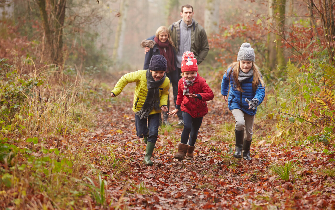 Eine Familie bei einem Spaziergang im Laubwald. Drei Kinder rennen voraus, die beiden Eltern laufen hinterher,