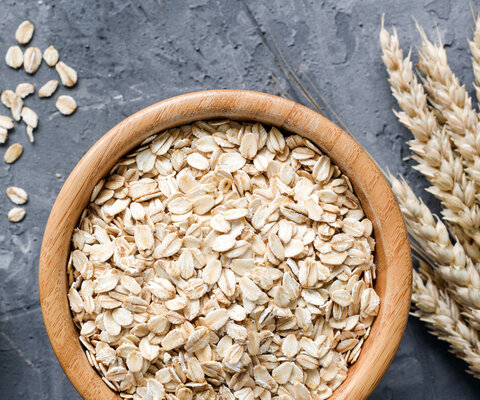 Haferflocken in einer Holzschüssel und goldene Getreideähren auf Steinhintergrund. 