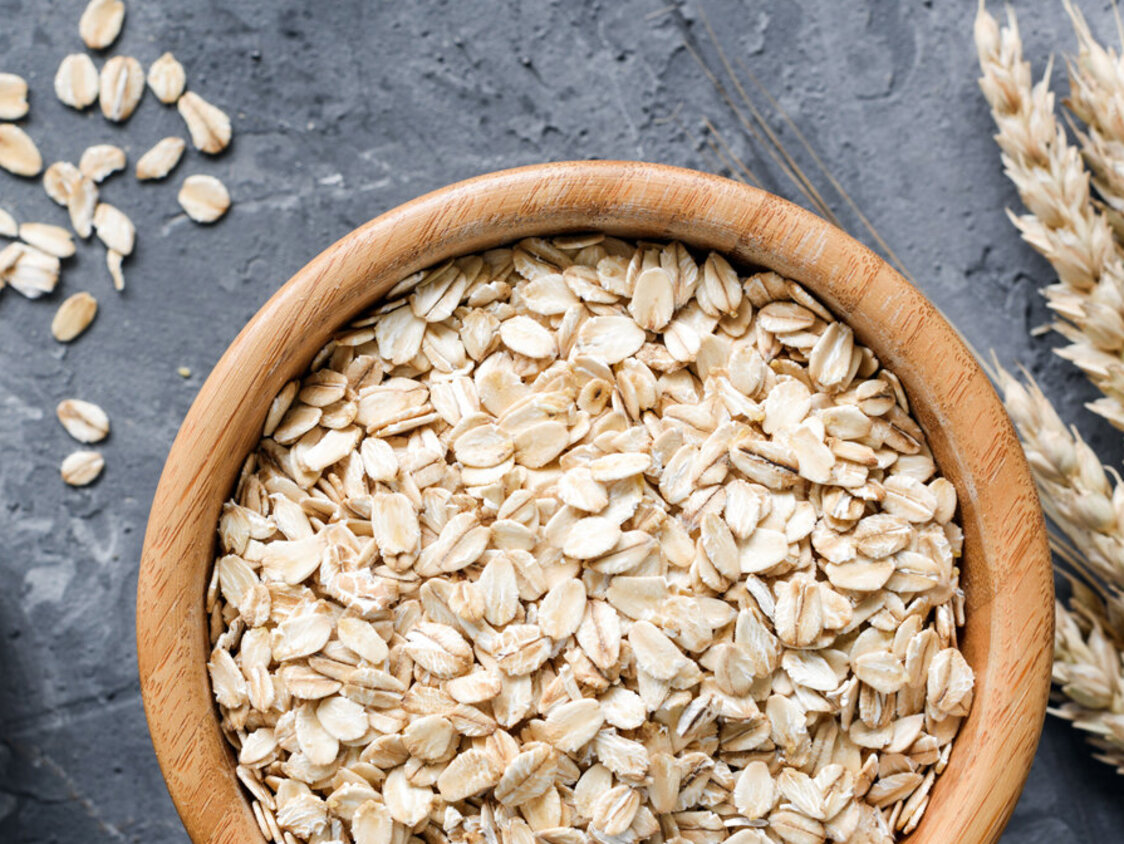 Haferflocken in einer Holzschüssel und goldene Getreideähren auf Steinhintergrund. 