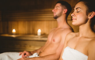 Ein Mann und eine Frau sitzen in weiße Handtücher gewickelt in einer Sauna. Beide haben die Augen geschlossen.
