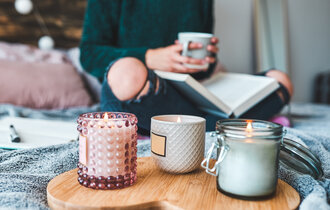 Drei brennende Kerzen stehen auf einem herzförmigen Holzbrett auf einer Decke. Im Hintergrund liest eine Frau ein Buch und hält eine Tasse in der Hand.