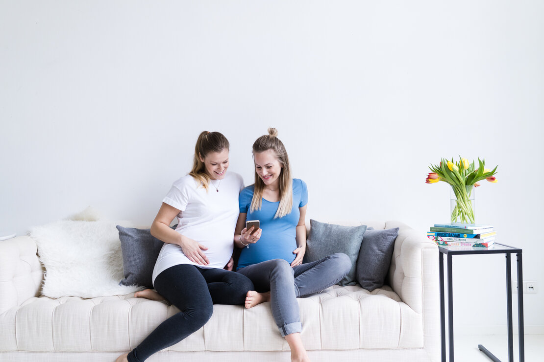 Zwei schwangere Frauen sitzen auf dem Sofa und schauen sich etwas auf dem Hady an.