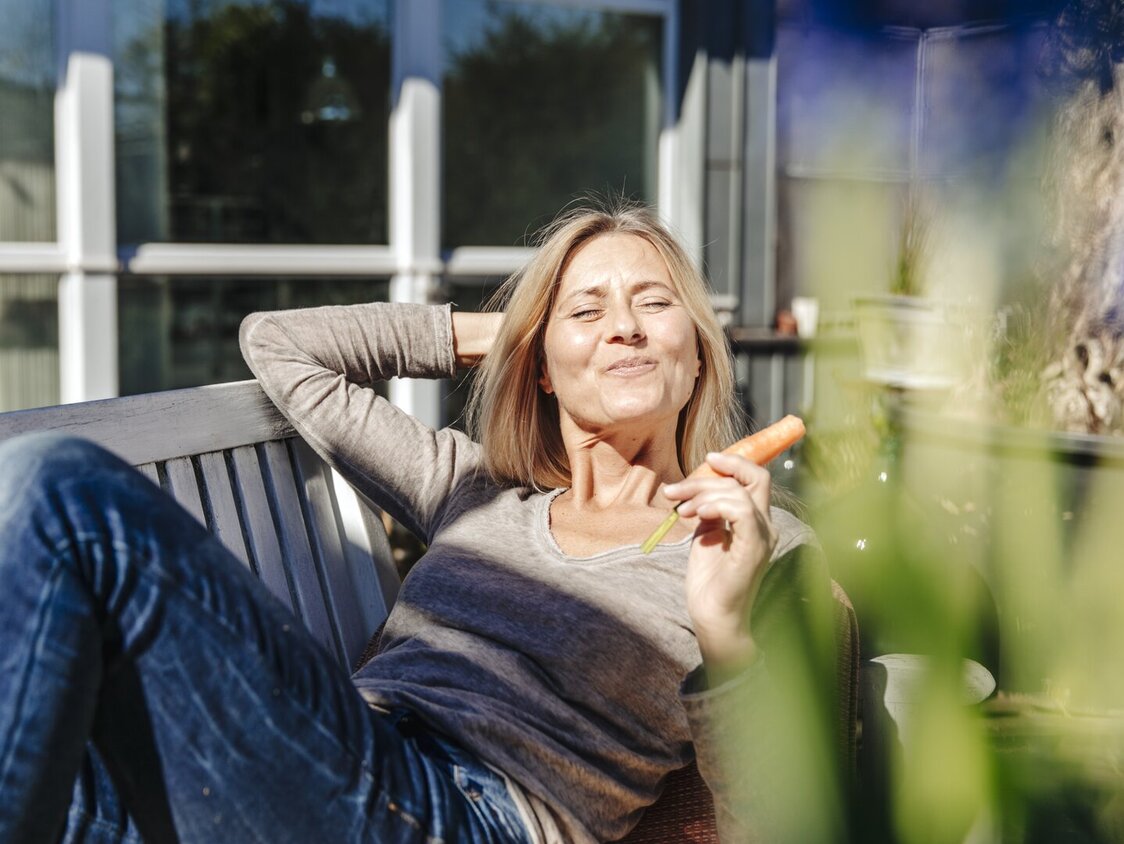 Frau legt auf einer Bank, hält eine Karotte in Ihrer Hand und hält Ihr Gesicht in die Sonne.