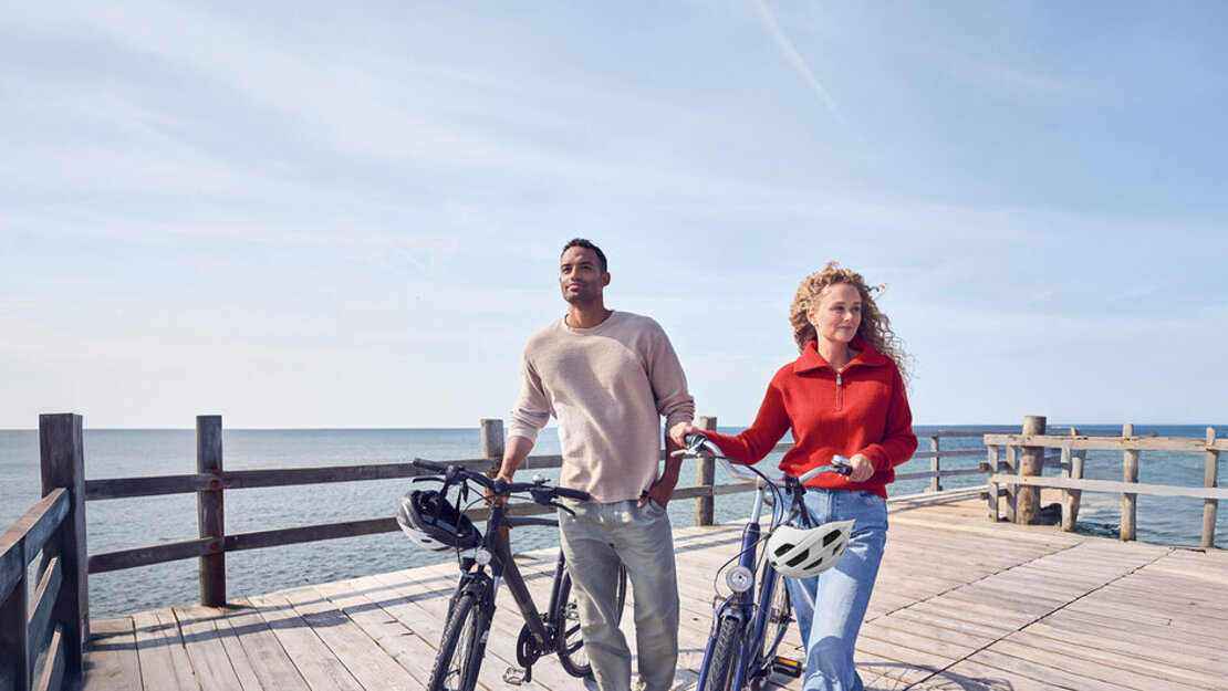 Eine Frau und ein Mann schieben auf einem Steg am Wasser ihre Farräder.