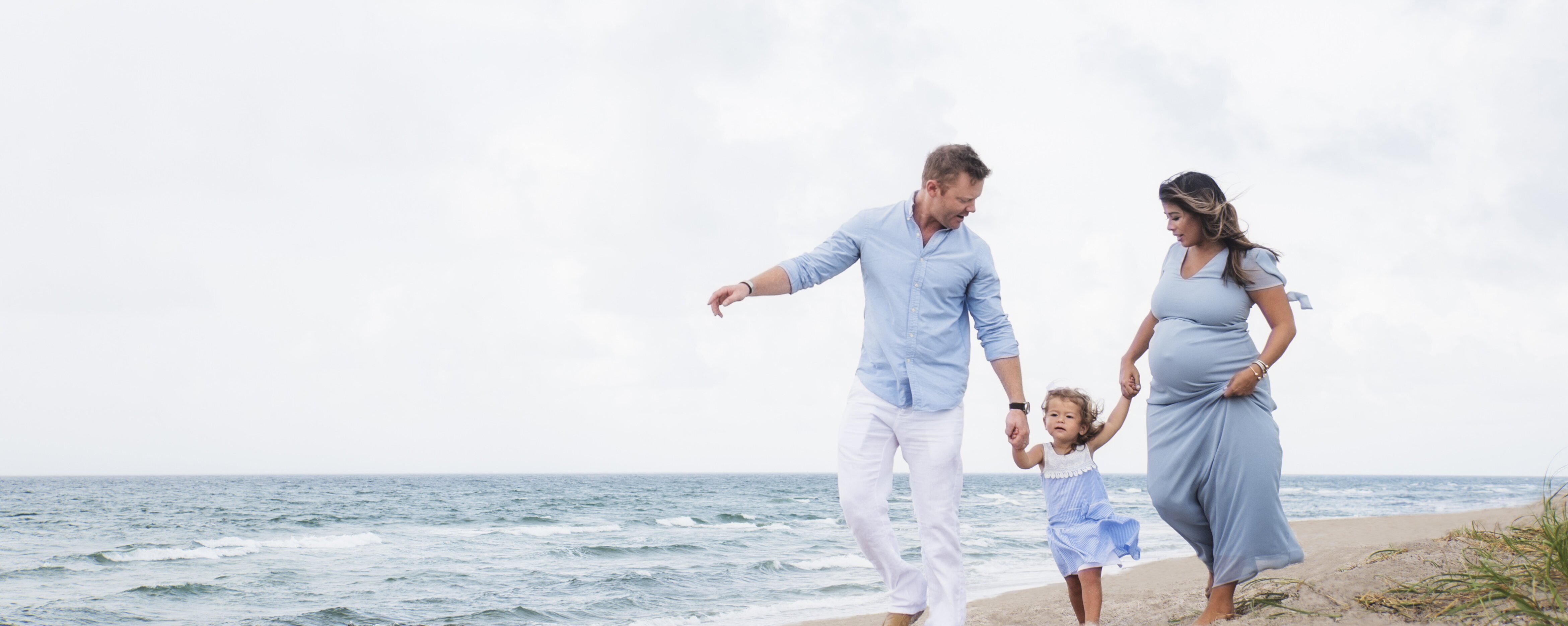 Eine Familie geht mit ihrem Kind am Strand spazieren. Die Eltern haben ihre Tochter an der Hand und die Mutter ist schwanger.