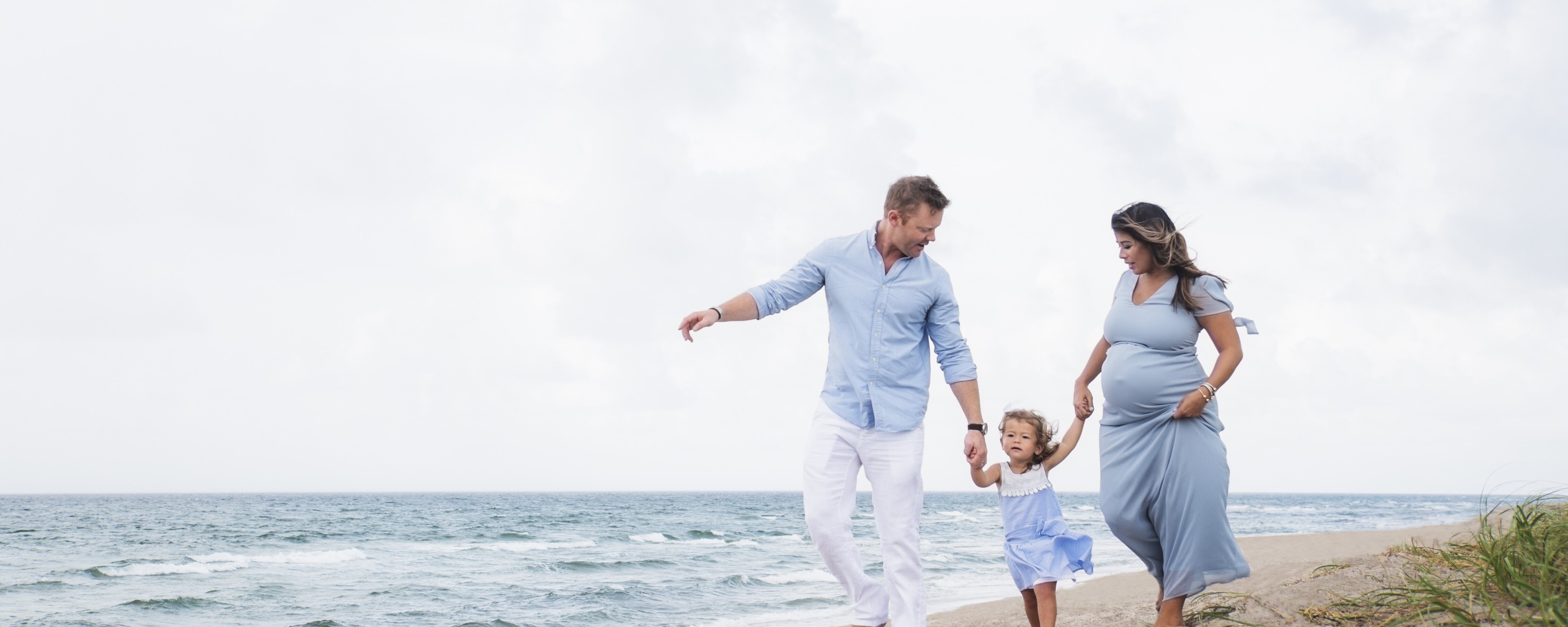 Eine Familie geht mit ihrem Kind am Strand spazieren. Die Eltern haben ihre Tochter an der Hand und die Mutter ist schwanger.