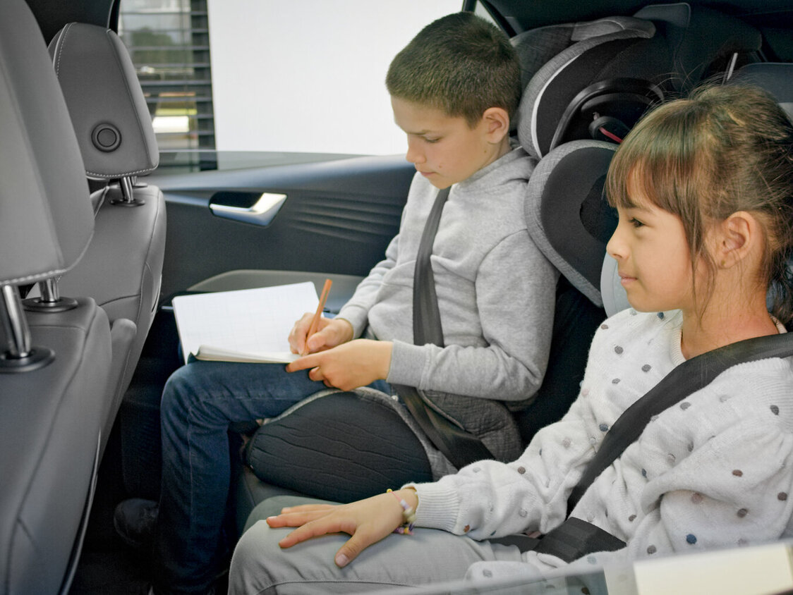 Ein Mädchen und ein Junge sitzen auf der Rückbank eines Autos und der Junge schreibt in ein Notizbuch.