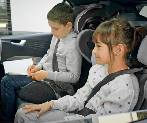 Ein Mädchen und ein Junge sitzen auf der Rückbank eines Autos und der Junge schreibt in ein Notizbuch.