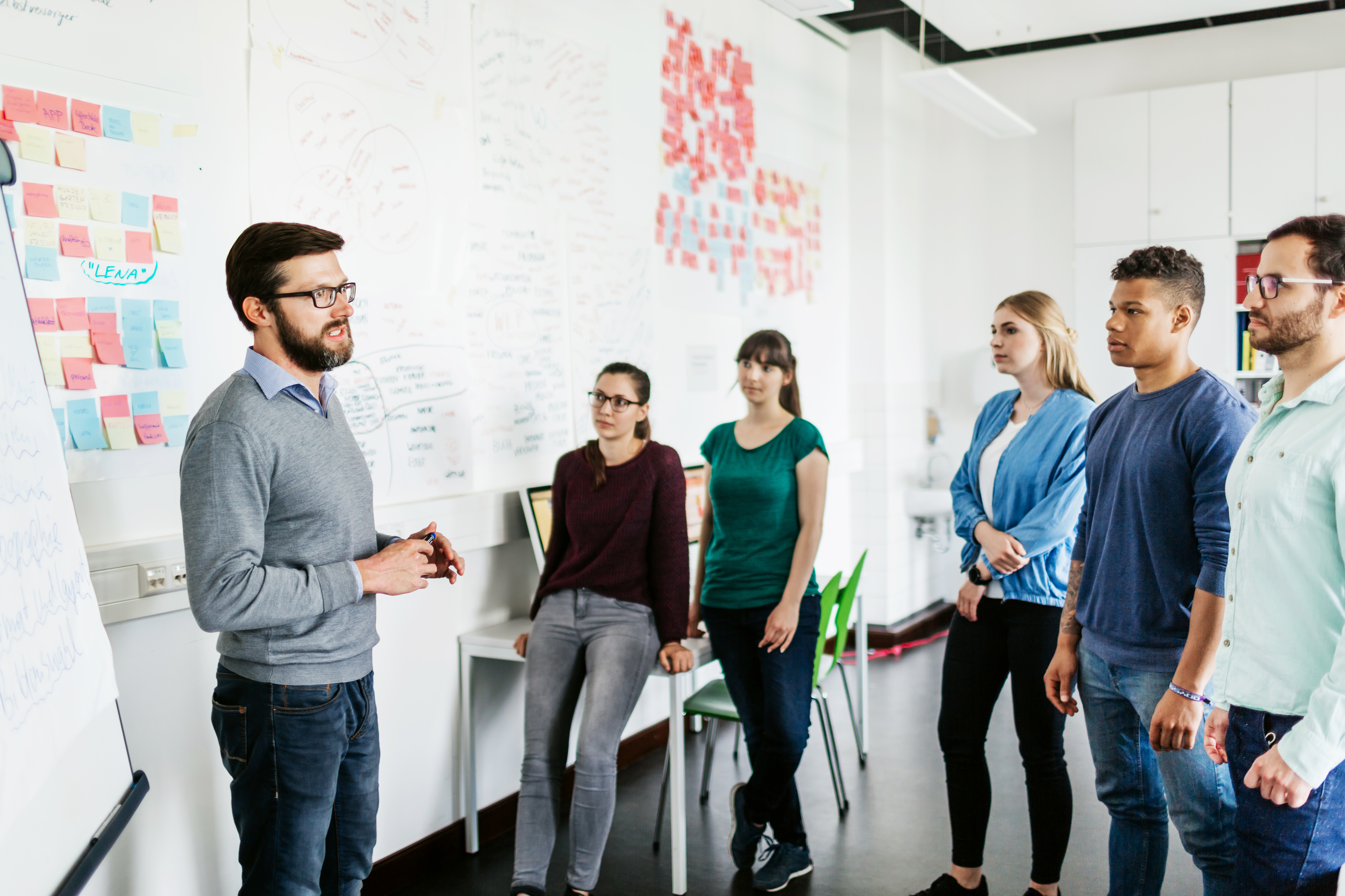 Ein Mann, welcher vor einer Gruppe junger Menschen steht und redet. Hinter ihm ist ein Whiteboard zu sehen. Alles spielt sich in einer Büroumgebung ab.